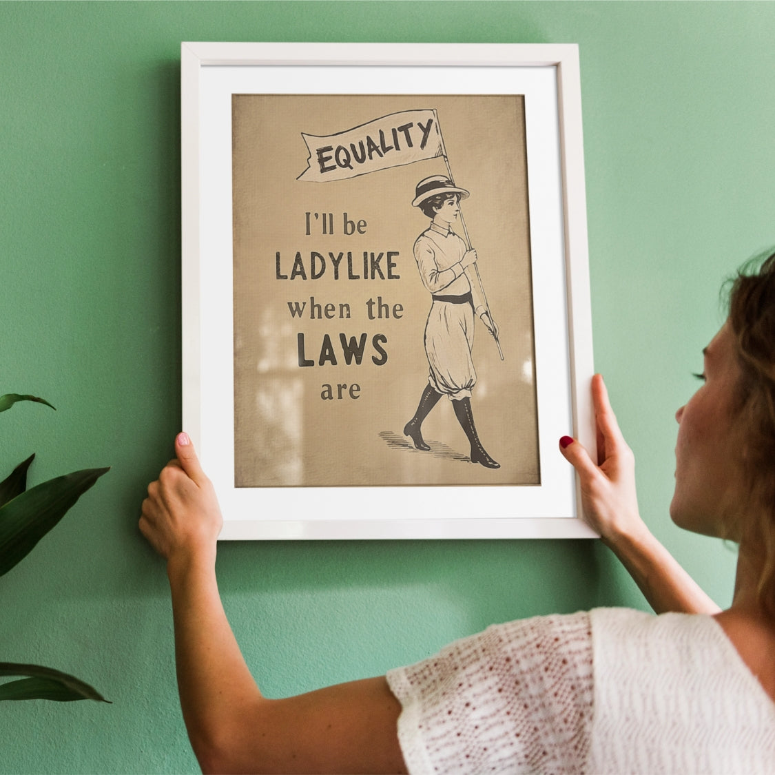 Person hanging a framed print with a vintage-style illustration and text on a green wall.