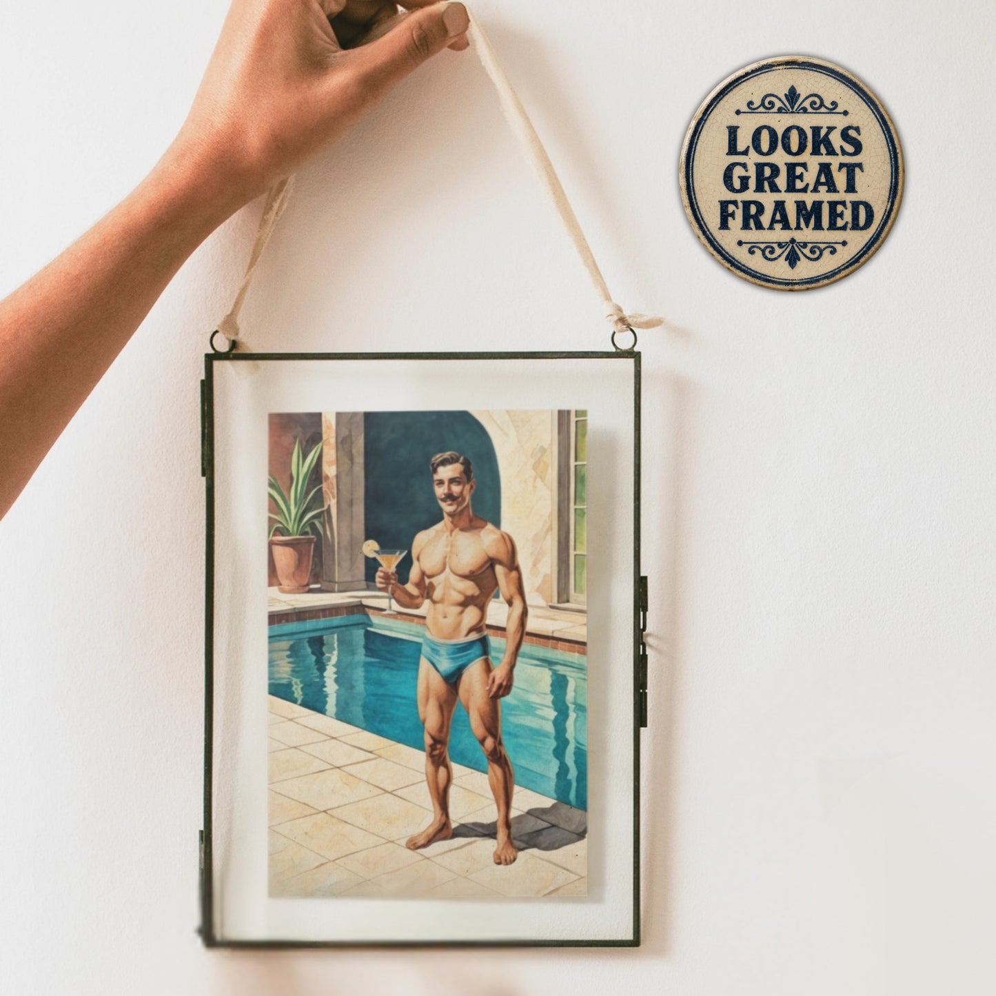 Framed greeting card artwork of a zaddy pinup man by a pool with a decorative sign reading 'Looks Great Framed' on a white wall.