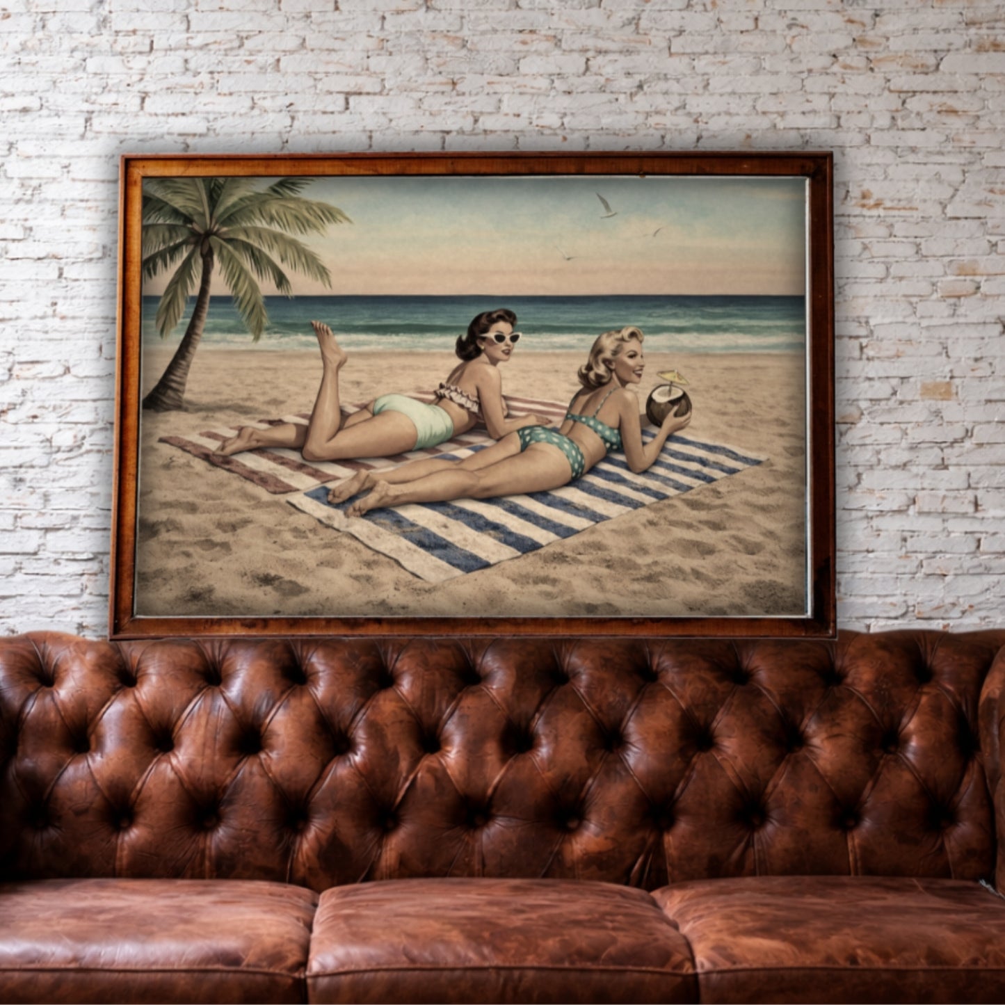 Framed artwork of two women on a beach with a palm tree, displayed above a brown leather sofa against a brick wall.