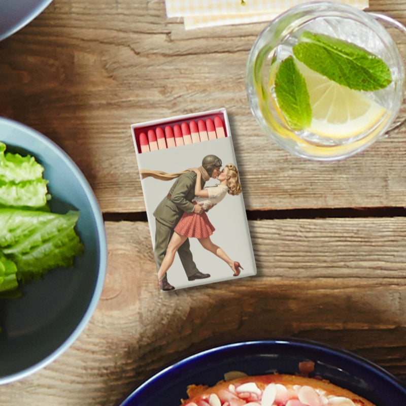 Matchbox with a design of a couple embracing, placed on a wooden surface with a glass of water and a bowl of salad.