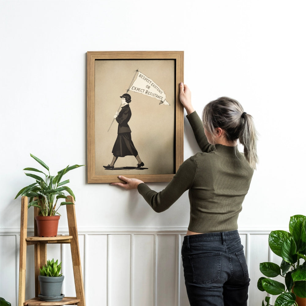 Person hanging a framed artwork titled: Respect Existence or Expect Resistance on a wall with plants around