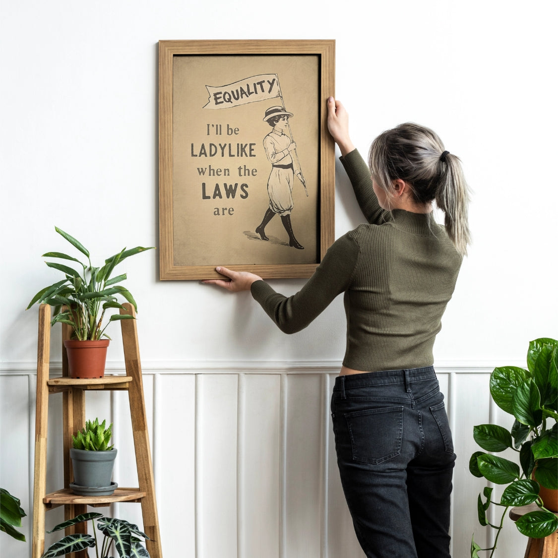 Person hanging a framed picture on a wall with plants around