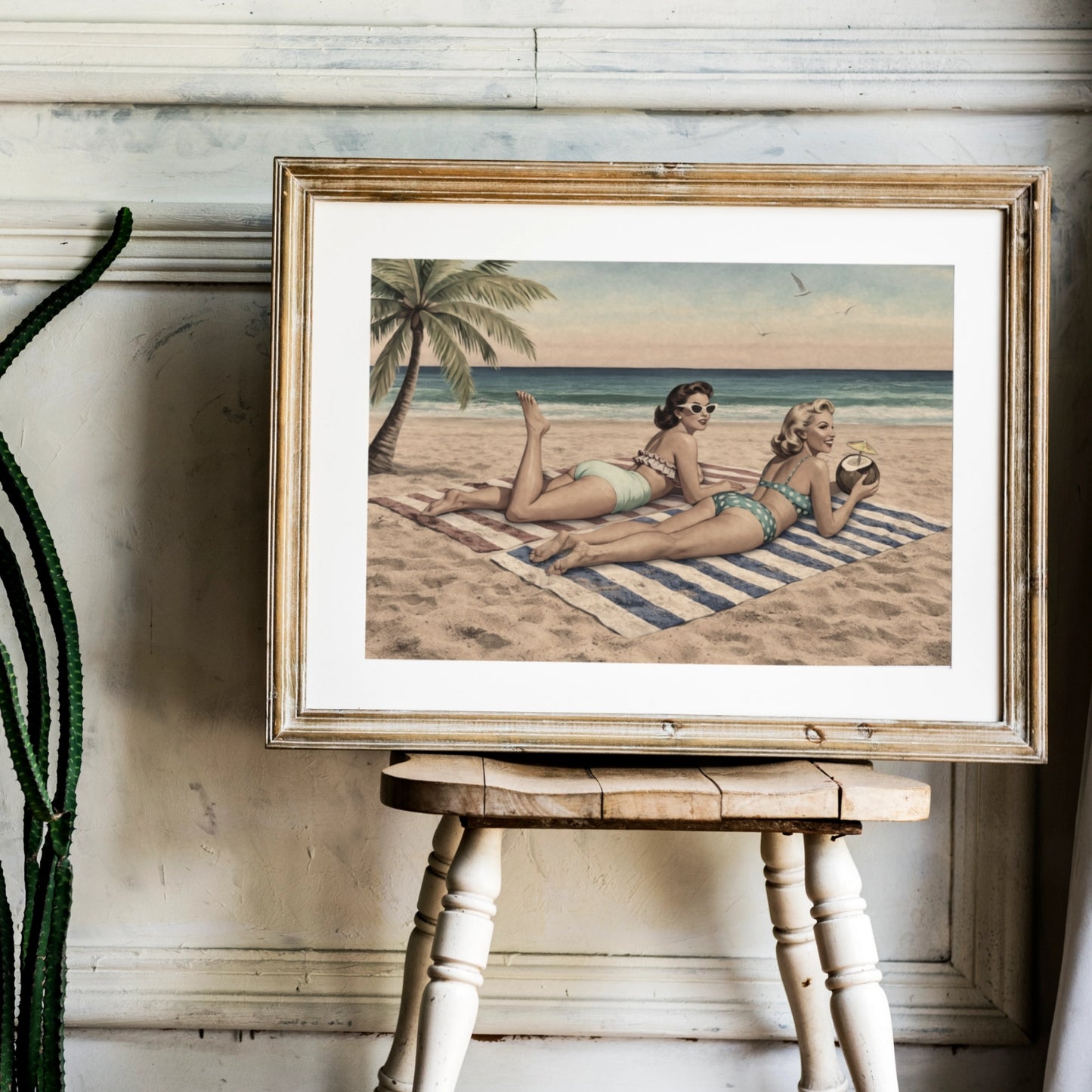 Framed artwork of two women on a beach with palm trees, displayed on a wooden stool.