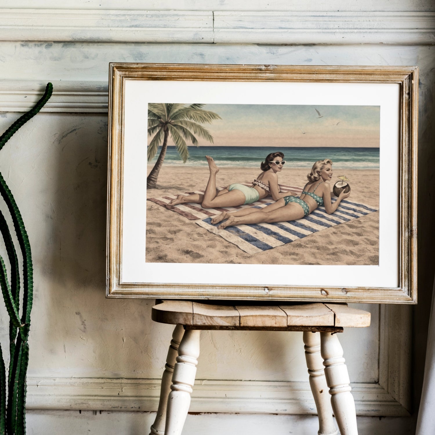 Framed artwork of two women on a beach with palm trees, displayed on a wooden stool.