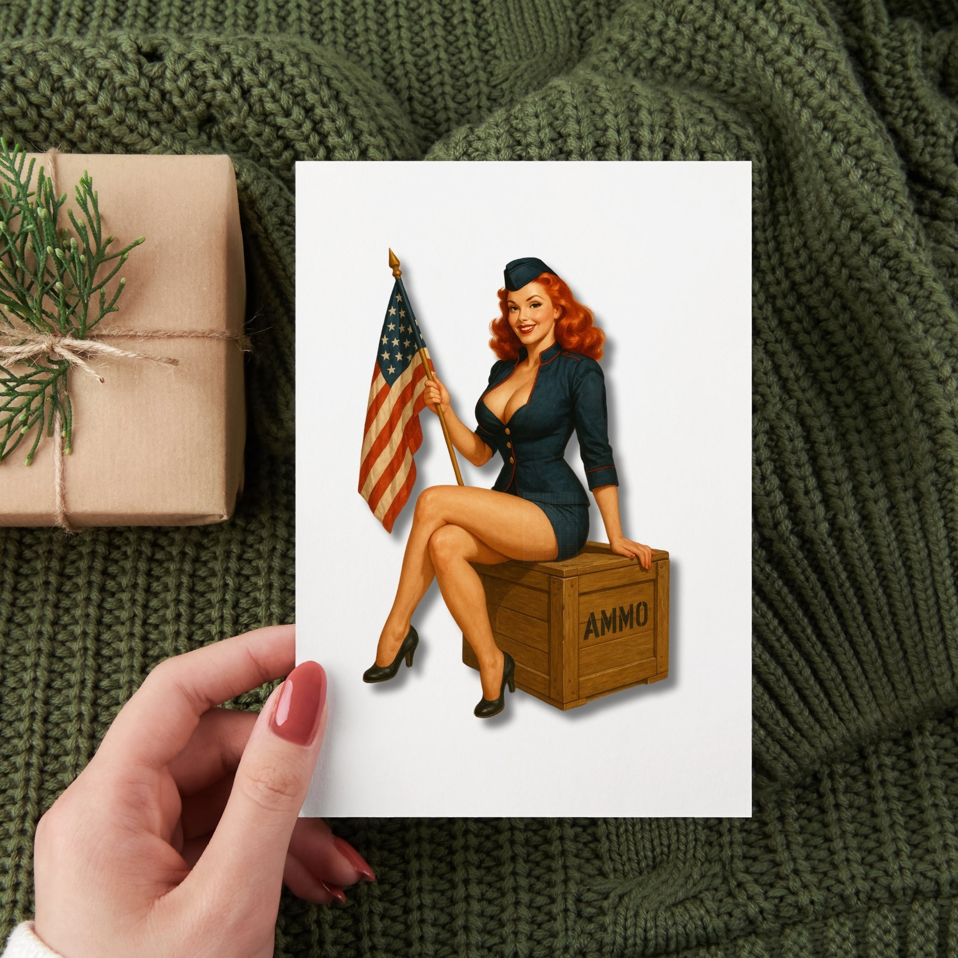 Hand holding a card with a vintage-style illustration of a woman holding an American flag and sitting on an 'AMMO' crate, on a textured green fabric background.