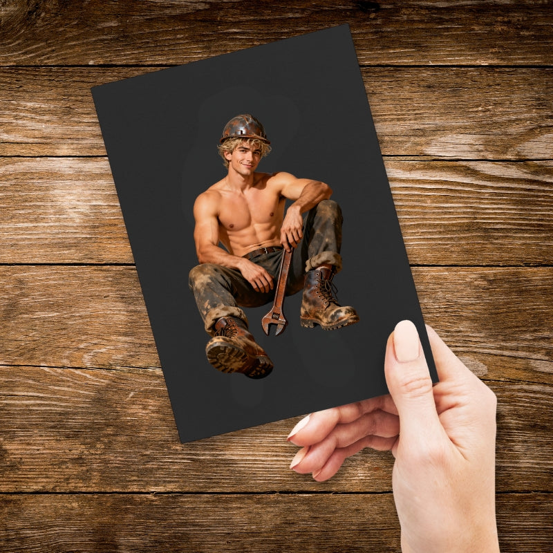 Hand holding a black card with a shirtless man in work attire and tools on a wooden surface